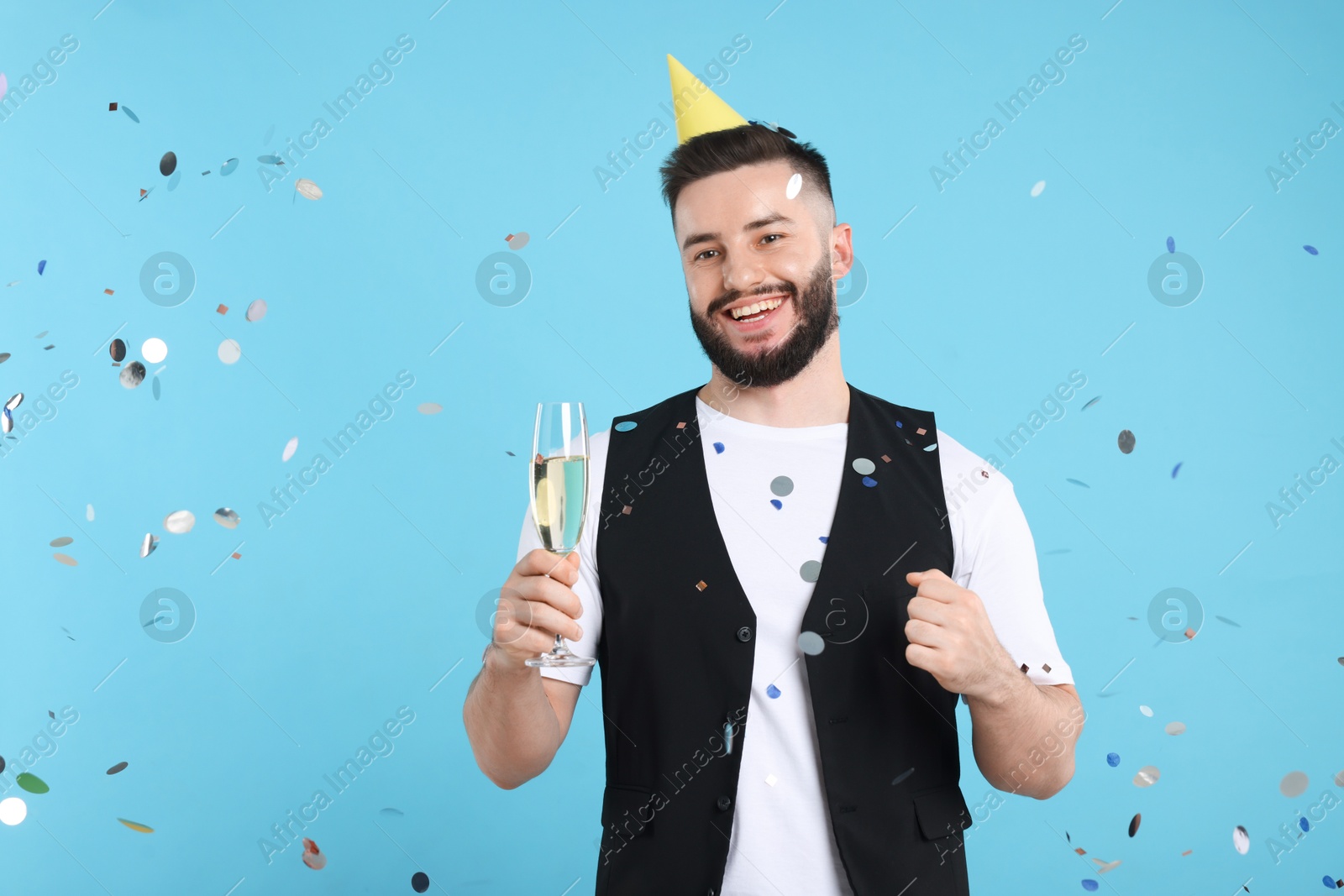 Happy Birthday. Handsome man with party hat and glass of champagne under confetti shower on light blue background, space for text Photo of Happy Birthday. Handsome man with party hat and glass of champagne under confetti shower on light blue background, space for text