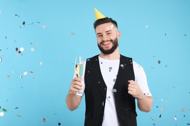 Photo of Happy Birthday. Handsome man with party hat and glass of champagne under confetti shower on light blue background, space for text