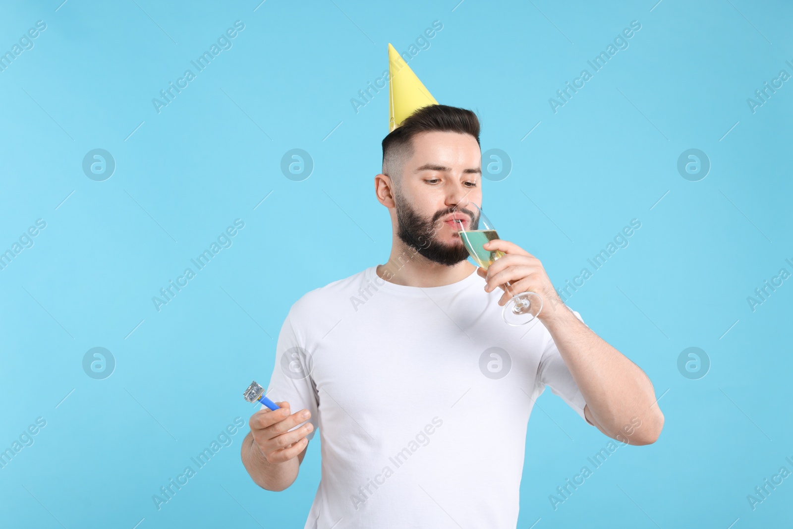 Happy Birthday. Handsome man with party hat, blower and glass of champagne on light blue background Photo of Happy Birthday. Handsome man with party hat, blower and glass of champagne on light blue background