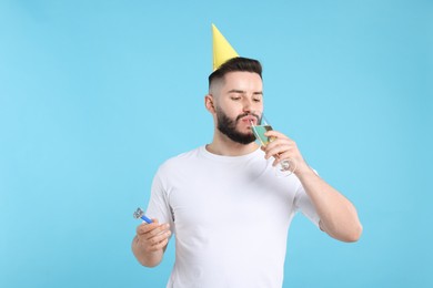 Photo of Happy Birthday. Handsome man with party hat, blower and glass of champagne on light blue background