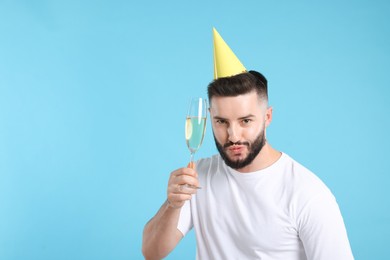 Photo of Happy Birthday. Handsome man with party hat and glass of champagne on light blue background, space for text