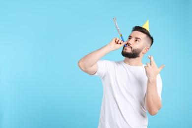 Photo of Happy Birthday. Handsome man with party hat and blower crossing fingers while making wish on light blue background, space for text