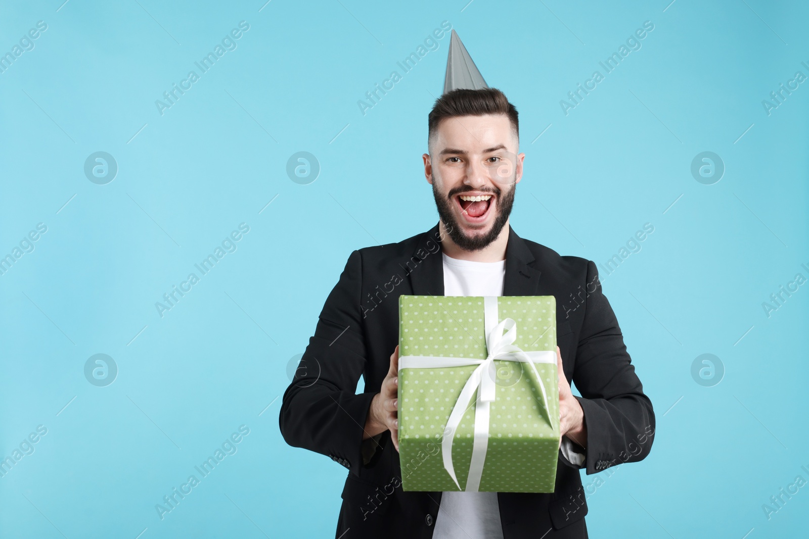 Happy Birthday. Handsome man with party hat and gift box on light blue background, space for text Photo of Happy Birthday. Handsome man with party hat and gift box on light blue background, space for text