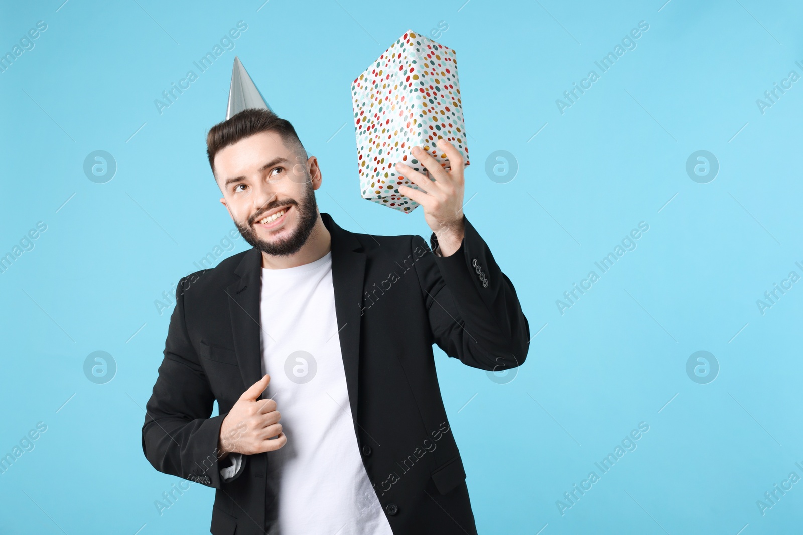 Happy Birthday. Handsome man with party hat and gift box on light blue background, space for text Photo of Happy Birthday. Handsome man with party hat and gift box on light blue background, space for text