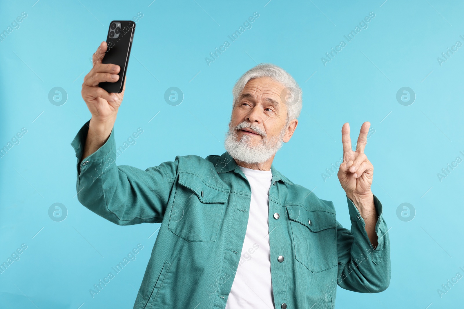 Elderly man showing peace sign while taking selfie on light blue background Photo of Elderly man showing peace sign while taking selfie on light blue background