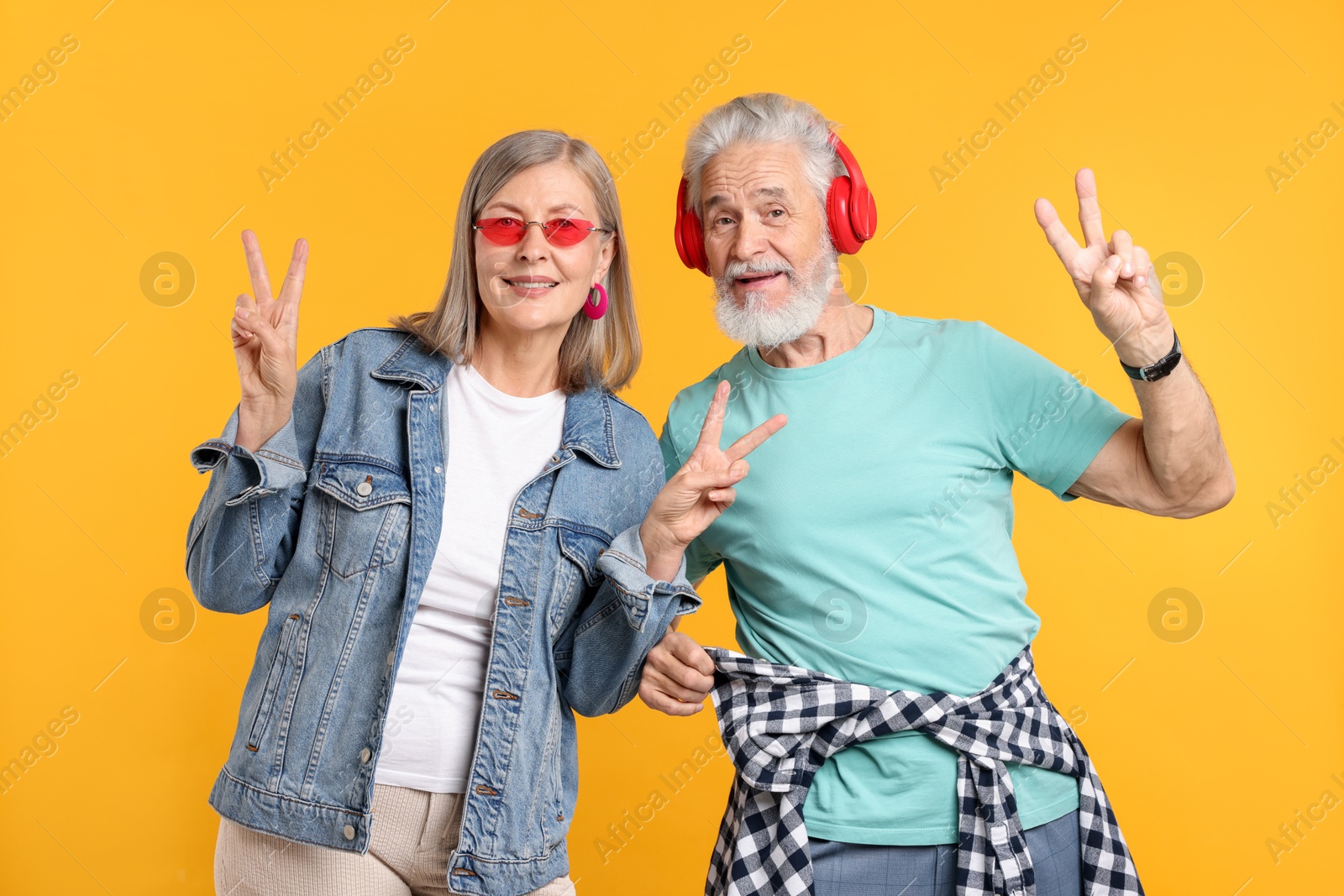 Senior couple in stylish outfits showing V-signs on orange background Photo of Senior couple in stylish outfits showing V-signs on orange background