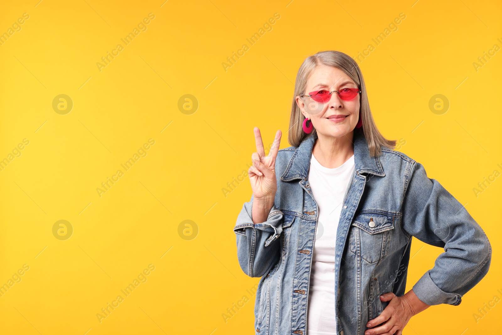 Senior woman in stylish outfit showing v-sign on orange background, space for text Photo of Senior woman in stylish outfit showing v-sign on orange background, space for text