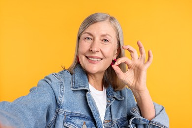 Photo of Senior woman in stylish outfit showing ok gesture while taking selfie on orange background