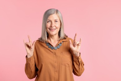 Photo of Senior woman in stylish outfit making rock gesture on pink background
