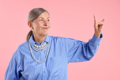 Photo of Senior woman wearing elegant pearl jewelry and making rock gesture on pink background