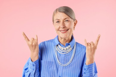 Photo of Senior woman wearing elegant pearl jewelry and making rock gesture on pink background