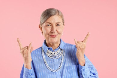 Photo of Senior woman wearing elegant pearl jewelry and making rock gesture on pink background