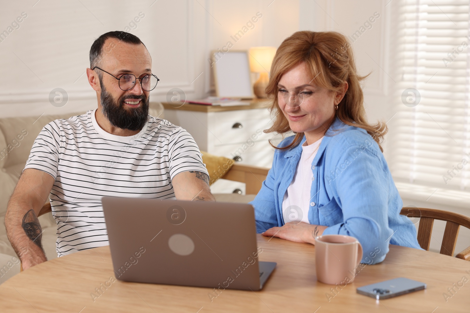 Lovely couple watching something on laptop at wooden table indoors Photo of Lovely couple watching something on laptop at wooden table indoors