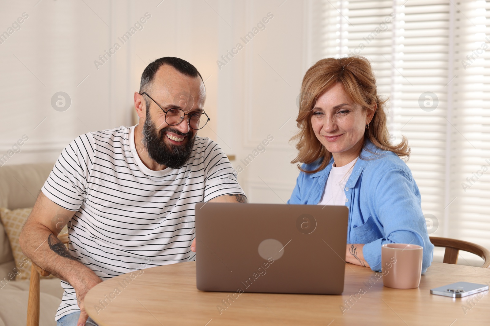 Lovely couple watching something on laptop at wooden table indoors Photo of Lovely couple watching something on laptop at wooden table indoors