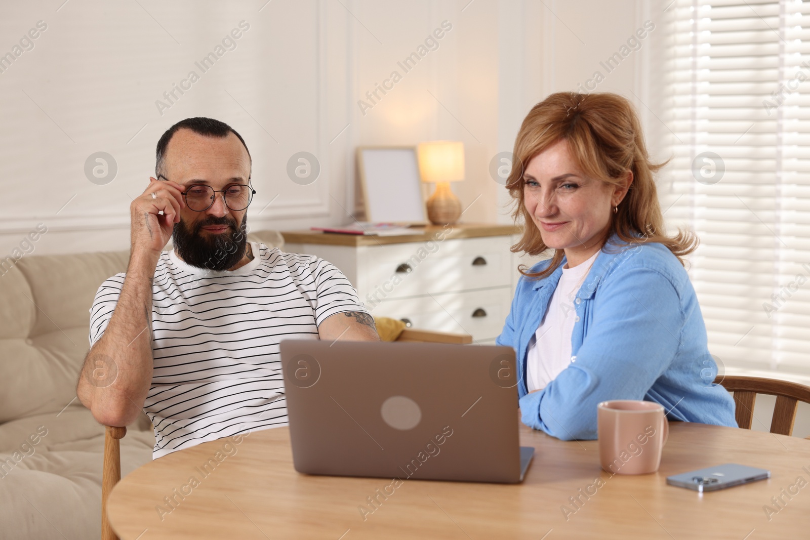 Lovely couple watching something on laptop at wooden table indoors Photo of Lovely couple watching something on laptop at wooden table indoors