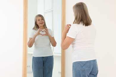 Photo of Beautiful senior woman making heart gesture near mirror indoors