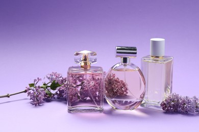 Photo of Bottles of perfumes and branches with lilac flowers on purple gradient background, closeup