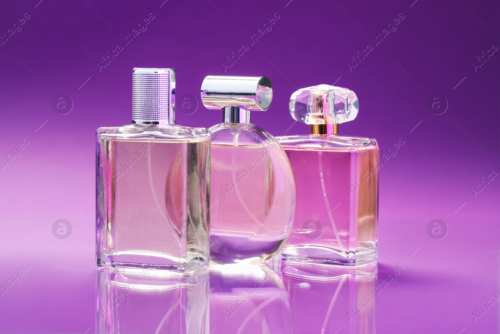 Photo of Bottles with perfumes on mirror surface against color gradient background, closeup
