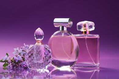 Photo of Bottles with perfumes and lilac flowers on mirror surface against color gradient background, closeup