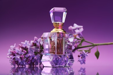 Photo of Bottle of perfume and branch with lilac flowers on mirror surface against color gradient background, closeup