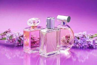 Photo of Bottles with perfumes and lilac flowers on mirror surface against color gradient background, closeup