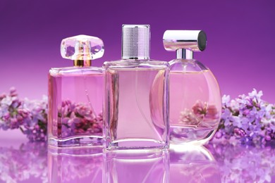 Photo of Bottles with perfumes and lilac flowers on mirror surface against color gradient background, closeup