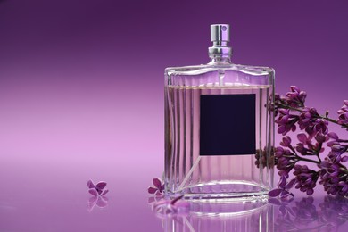 Photo of Bottle with perfume and lilac flowers on mirror surface against color gradient background, closeup. Space for text