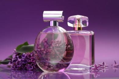 Photo of Bottles with perfumes and lilac flowers on mirror surface against color gradient background, closeup