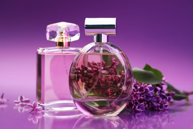 Photo of Bottles with perfumes and lilac flowers on mirror surface against color gradient background, closeup