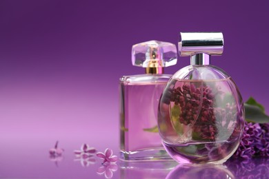 Photo of Bottles with perfumes and lilac flowers on mirror surface against color gradient background, closeup. Space for text