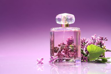 Photo of Bottle with perfume and lilac flowers on mirror surface against color gradient background, closeup. Space for text