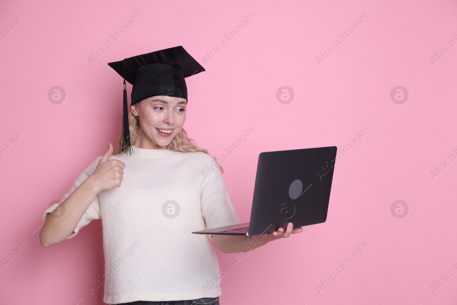 Happy student with laptop showing thumbs up after graduation on pink background Photo of Happy student with laptop showing thumbs up after graduation on pink background