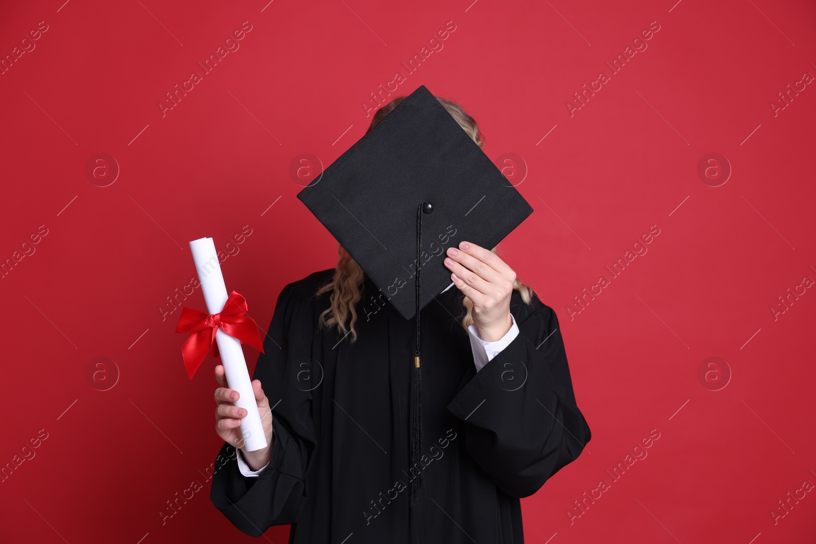 Student with diploma hiding behind mortarboard after graduation on red background Photo of Student with diploma hiding behind mortarboard after graduation on red background