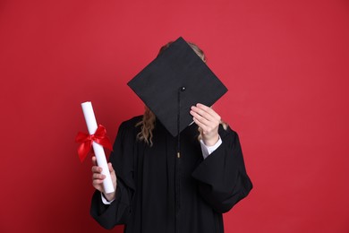 Photo of Student with diploma hiding behind mortarboard after graduation on red background