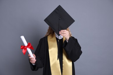 Photo of Student with diploma hiding behind mortarboard after graduation on grey background