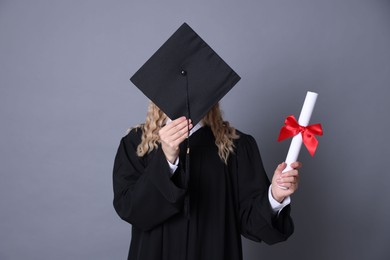 Photo of Student with diploma hiding behind mortarboard after graduation on grey background