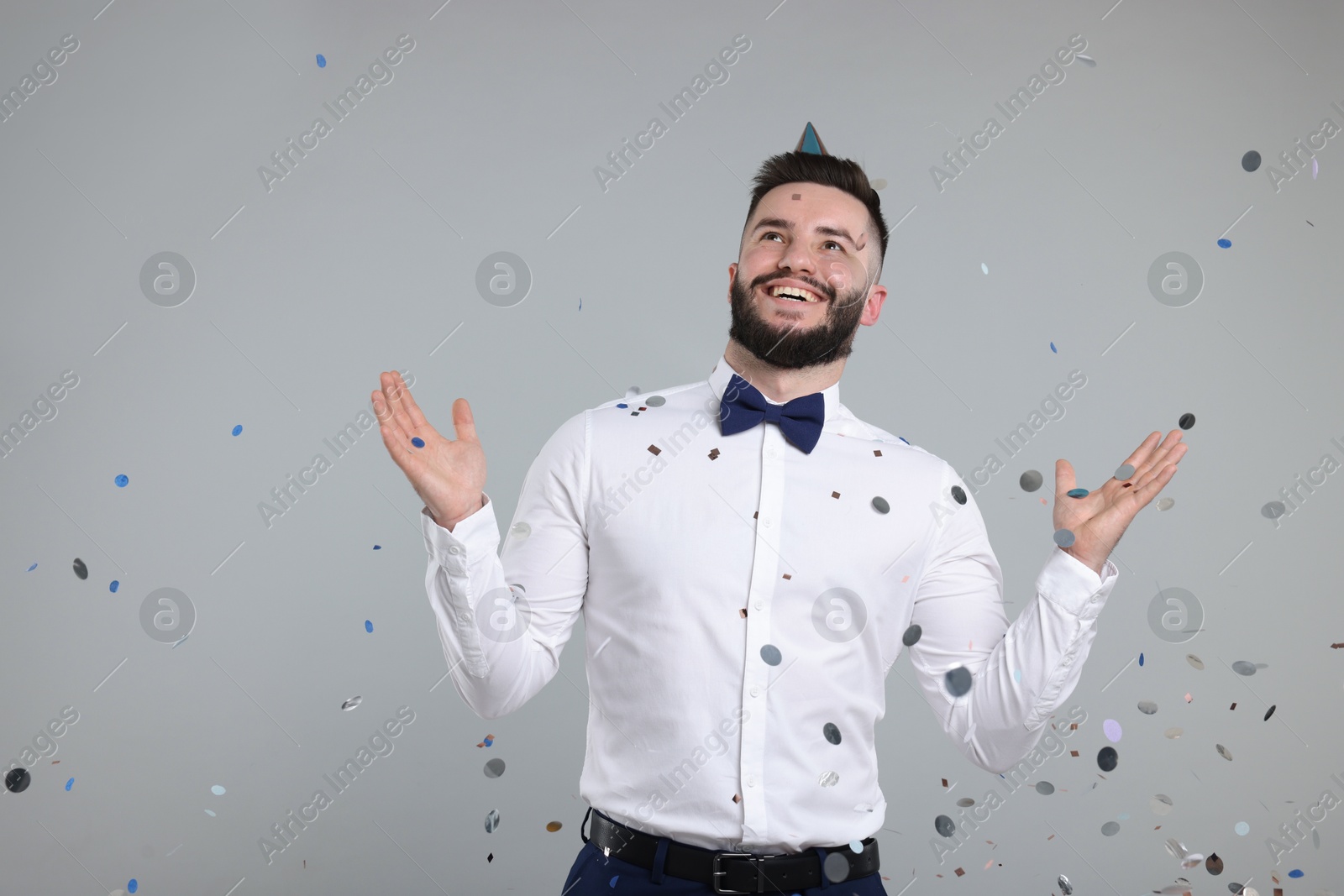 Happy Birthday. Handsome man with party hat under confetti shower on grey background, space for text Photo of Happy Birthday. Handsome man with party hat under confetti shower on grey background, space for text
