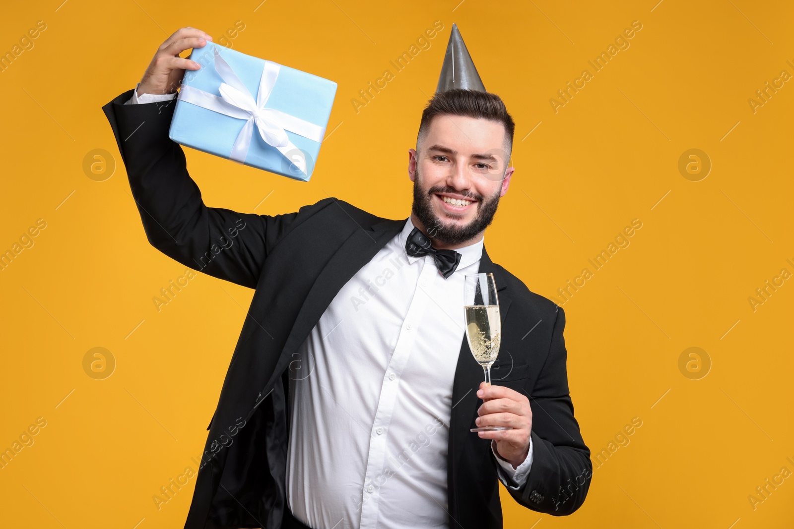 Happy Birthday. Handsome man with party hat, glass of champagne and gift box on orange background Photo of Happy Birthday. Handsome man with party hat, glass of champagne and gift box on orange background