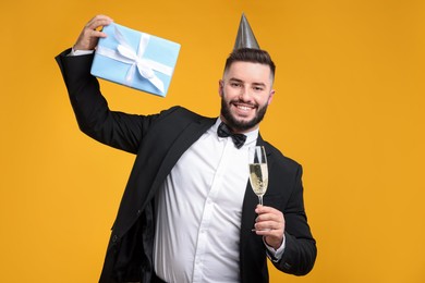 Photo of Happy Birthday. Handsome man with party hat, glass of champagne and gift box on orange background
