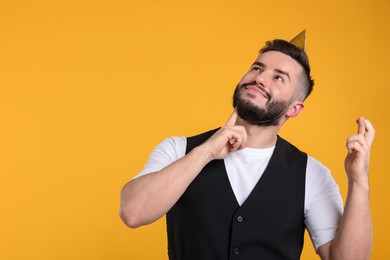 Photo of Happy Birthday. Handsome man with party hat crossing fingers while making wish on orange background, space for text