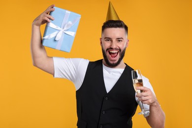 Photo of Happy Birthday. Handsome man with party hat, glass of champagne and gift box on orange background
