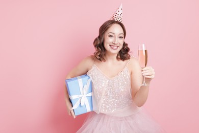 Photo of Birthday celebration. Woman with glass of sparkling wine and gift on pink background