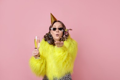 Photo of Birthday celebration. Woman with glass of sparkling wine blowing confetti on pink background