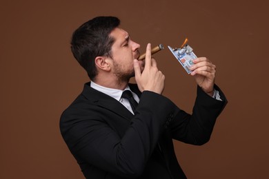 Photo of Greedy banker lighting cigar with dollar banknote on brown background