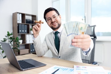 Photo of Happy greedy banker with dollar banknotes and cigar at table in office, selective focus