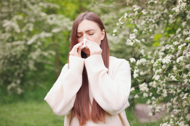 Photo of Woman with paper tissue suffering from seasonal pollen allergy near blossoming shrub on spring day