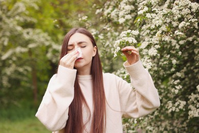 Photo of Woman with paper tissue suffering from seasonal pollen allergy near blossoming shrub on spring day