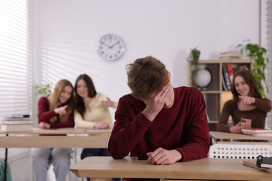 Photo of Cyber bullying. Upset teenage boy with smartphone and schoolmates pointing at him in classroom, selective focus