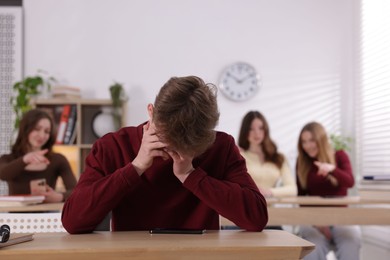 Photo of Cyber bullying. Upset teenage boy with smartphone and schoolmates pointing at him in classroom, selective focus