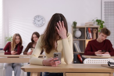 Photo of Cyber bullying. Upset teenage girl with smartphone at desk in classroom